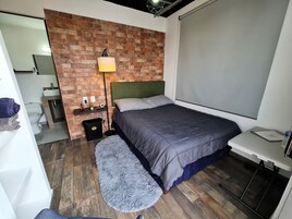 A bedroom with a brick wall, a bed with a green headboard, a bedside table, a lamp, and a view of the bathroom.