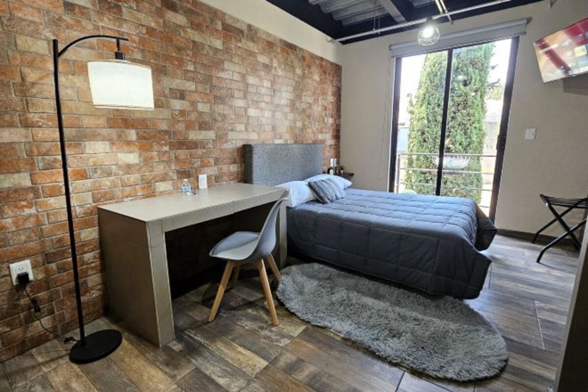Een slaapkamer met een bakstenen muur, een bed, een bureau met stoel, een groot raam en een televisie aan de muur.