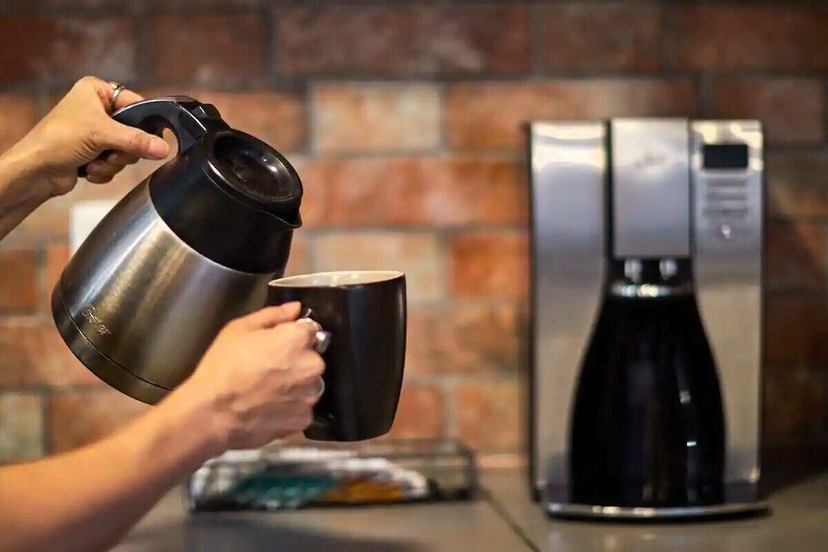 A person pouring coffee from a stainless steel pot into a black mug.