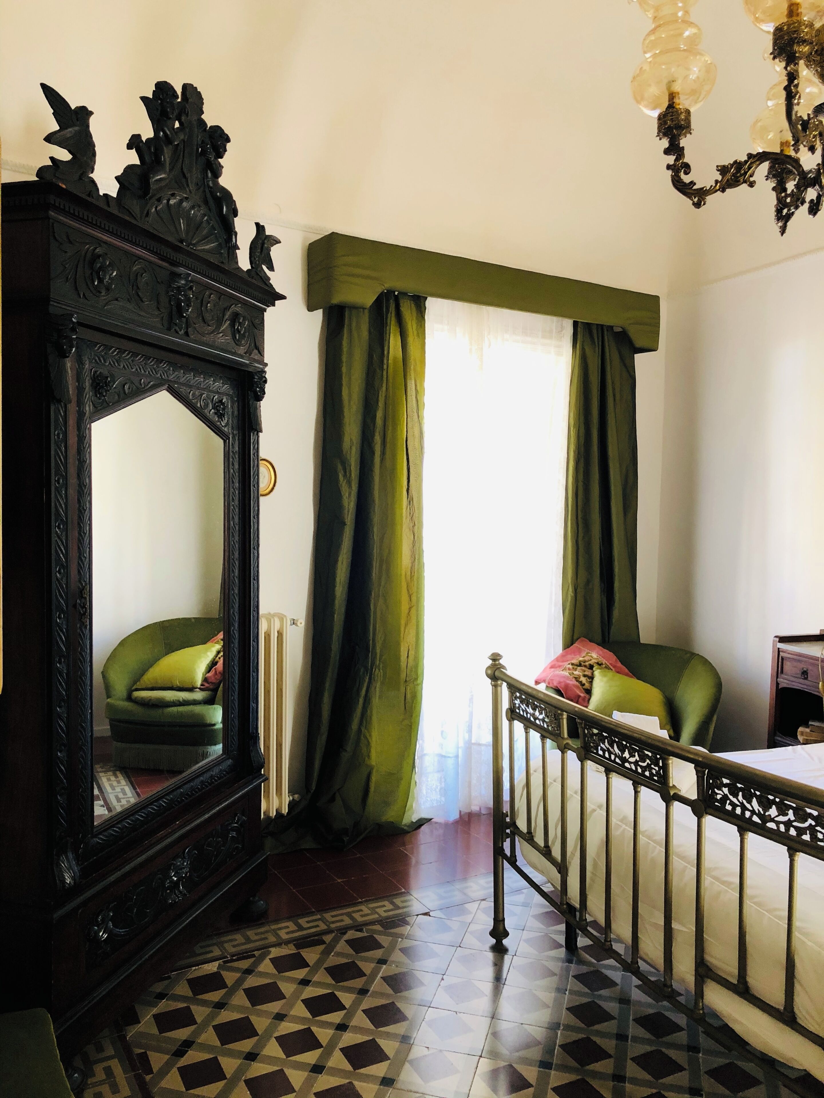A room with a large ornate mirror, a green upholstered chair, a bed with a metal headboard, and a patterned floor.