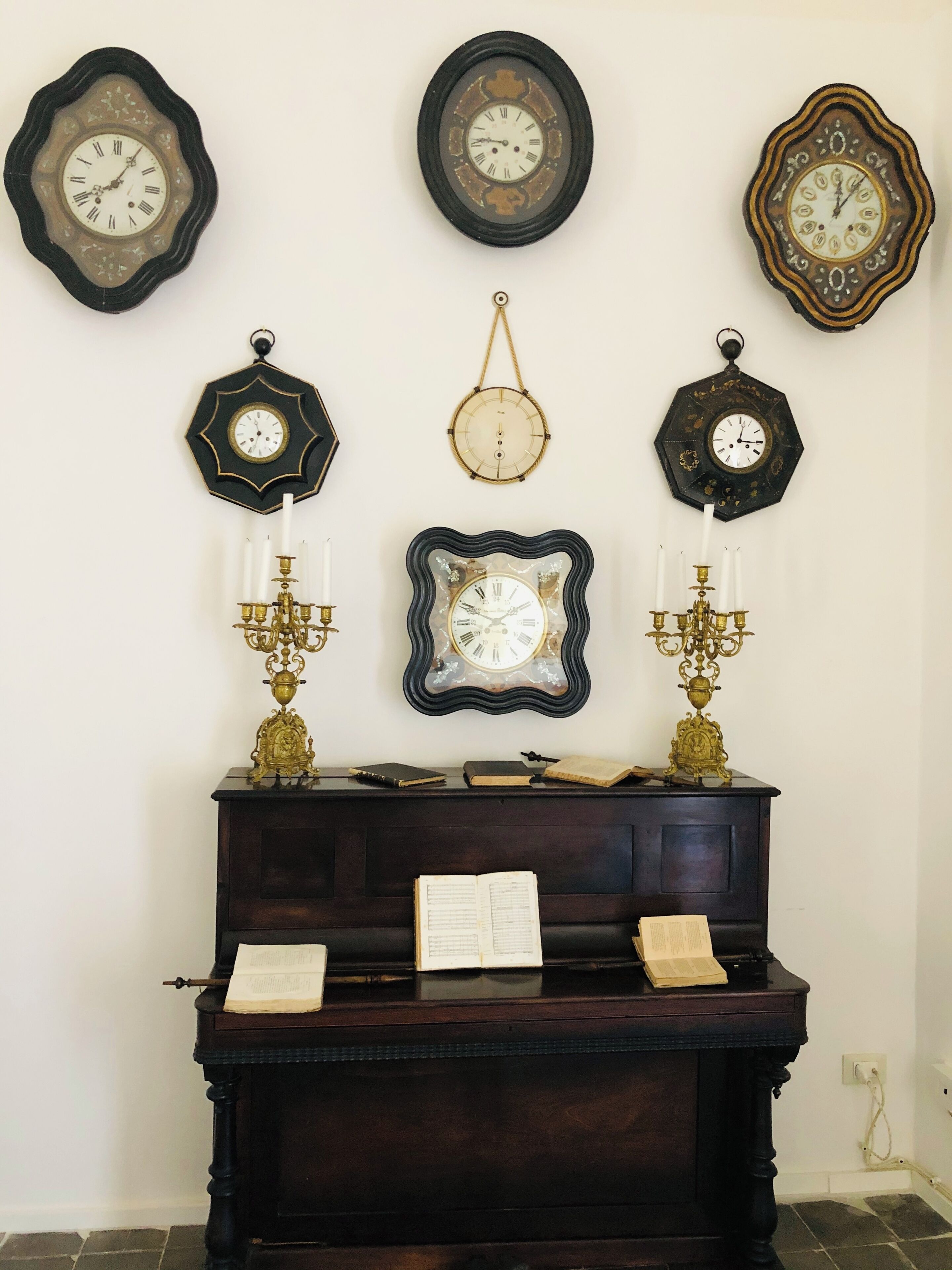 Una habitación con un piano de madera oscura, varios relojes fijos en la pared y dos candelabros elaborados.