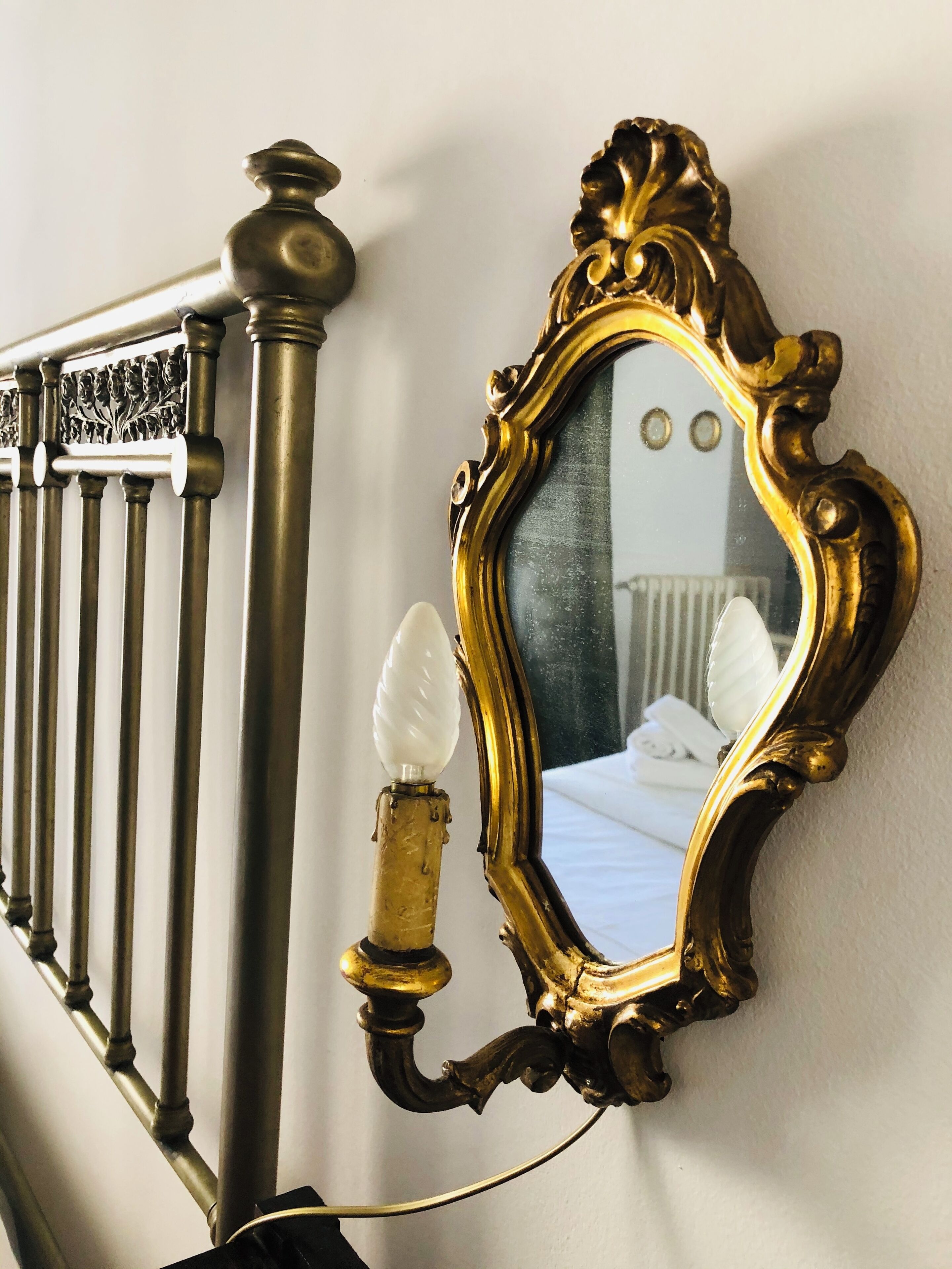 A bed with white linens, a gold-framed mirror, and a white light fixture on a wall.
