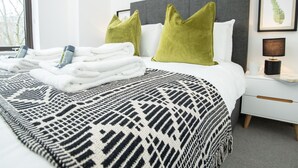 A neatly made bed with folded towels and toiletries, a patterned throw blanket, and a nightstand with a lamp and a framed picture. - Exeter Cathedral Two-bedroom Stay (Exeter)