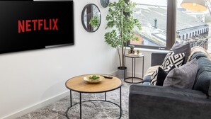 A living room with a flat-screen TV displaying "NETFLIX", a round coffee table, a sofa with cushions, a small side table with a plant, and a window with a view of buildings. - Exeter Cathedral Two-bedroom Stay (Exeter)