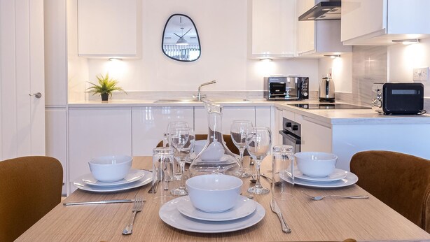 A modern kitchen with white cabinetry, a built-in oven, a microwave, and a toaster. There is a dining table set with white plates, bowls, and cutlery. - Victoria Square Two-bed Accommodation by Belvilla (Birmingham)