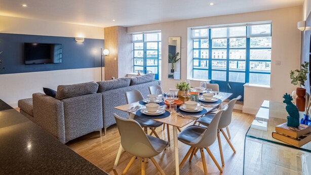 A modern living room with a sofa, dining table, and chairs. There is a flat-screen TV mounted on the wall, a window with a view of buildings, and a potted plant on the table.