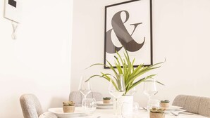 Una mesa redonda de comedor con platos blancos, cubiertos y copas de vino transparentes. Hay pequeñas macetas con plantas y un cuadro en la pared.
