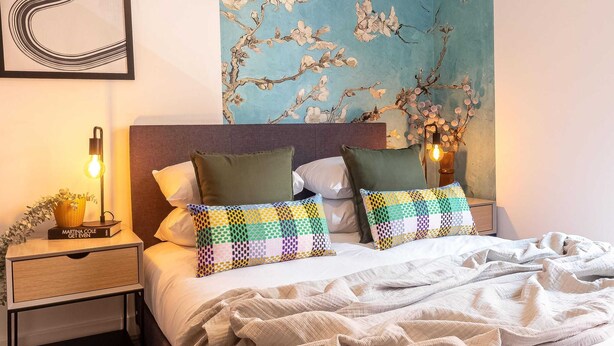 A neatly made bed with patterned pillows, a bedside lamp, and a wall art of branches with flowers. - One Bed Apartment Gem Steps to City by Belvilla (Birmingham)