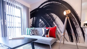 A modern living room with a grey sofa, patterned cushions, a black coffee table, and a large abstract painting on the wall. - One Bed Apartment Gem Steps to City by Belvilla (Birmingham)