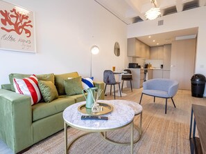 Un salon moderne avec un canapé vert, une table basse ronde et un fauteuil gris. On aperçoit une cuisine à l’arrière-plan, avec une table à manger et des chaises.