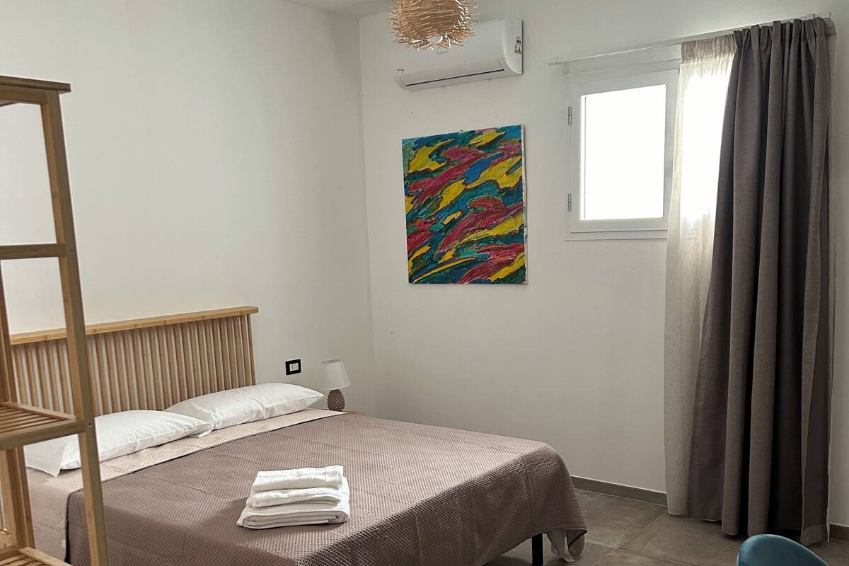 A bedroom with a bed, a wooden ladder, a desk, a chair, a window with curtains, an air conditioner, and a colorful painting on the wall.