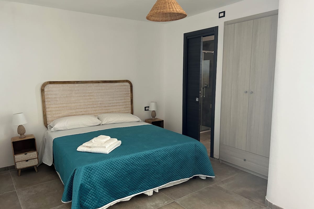 A neatly made bed with a teal bedspread and white pillows, a wooden headboard, a bedside lamp, and a small wicker basket on a bedside table.