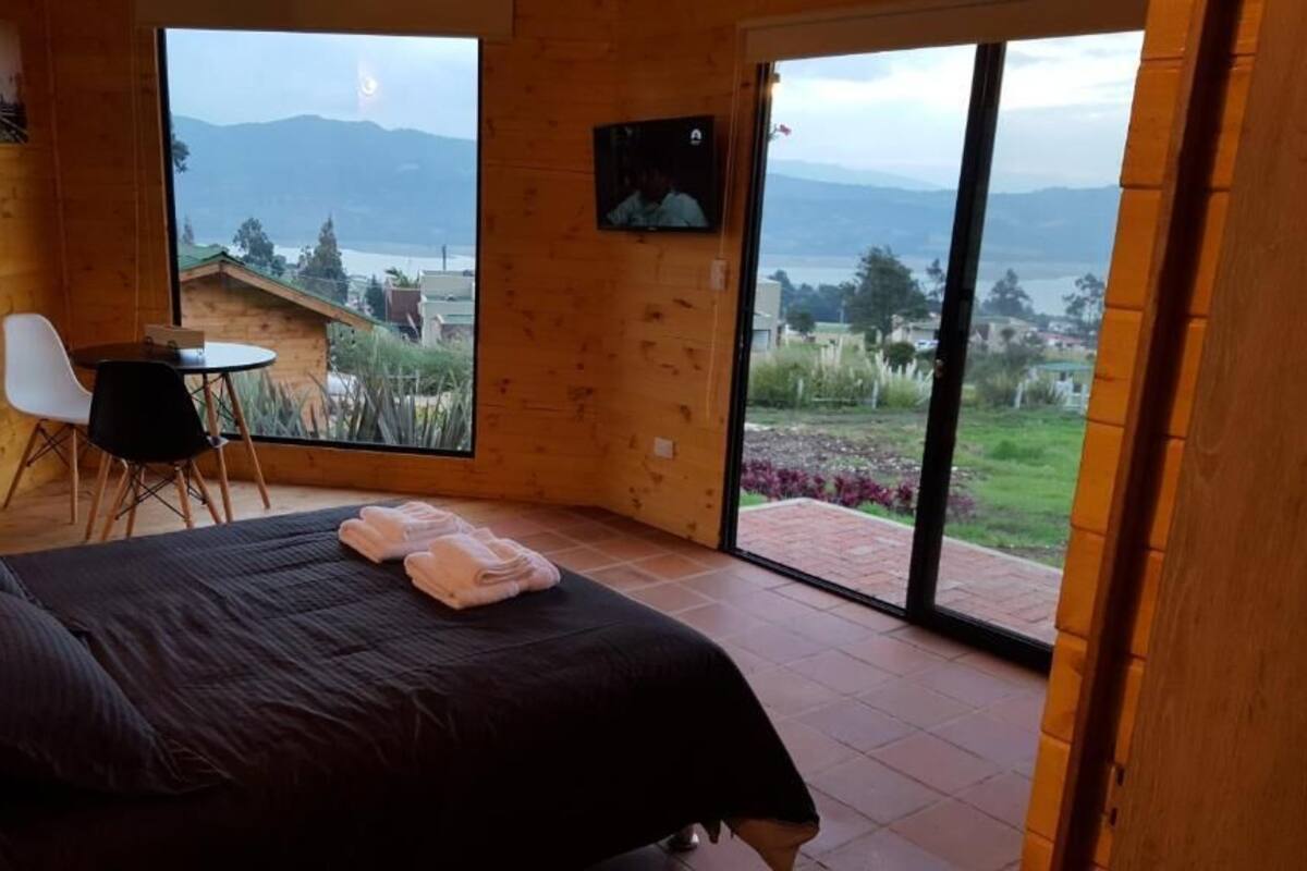 A wooden room with a bed, a small table, and a chair. There is a large window with a view of a mountain and a body of water.