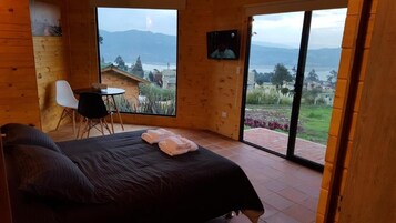Una habitación de madera con una cama, una mesita y una silla. Se aprecia una ventana grande con vistas a una montaña y una masa de agua.