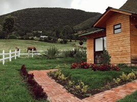 Una cabaña de madera con un sendero de ladrillo que lleva a la entrada, rodeada por una cerca blanca y un campo cubierto de césped con una vaca al fondo.