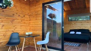 Una cabaña de madera con puerta de vidrio que da a un balcón, dos sillas, una mesita y una cama con almohadas.