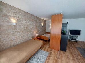 Quarto moderno com parede de tijolos, cama de madeira, mesa de cabeceira com luminária, televisão sobre um suporte e uma pequena planta.