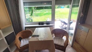A room with a table and two wicker chairs, a window with a view of a garden, and a sliding glass door leading outside. - 5 Minuten von Frankfurt, Ruhige Ferienwohnung/ Monteurzimmer mit Terrasse (Bad Vilbel)