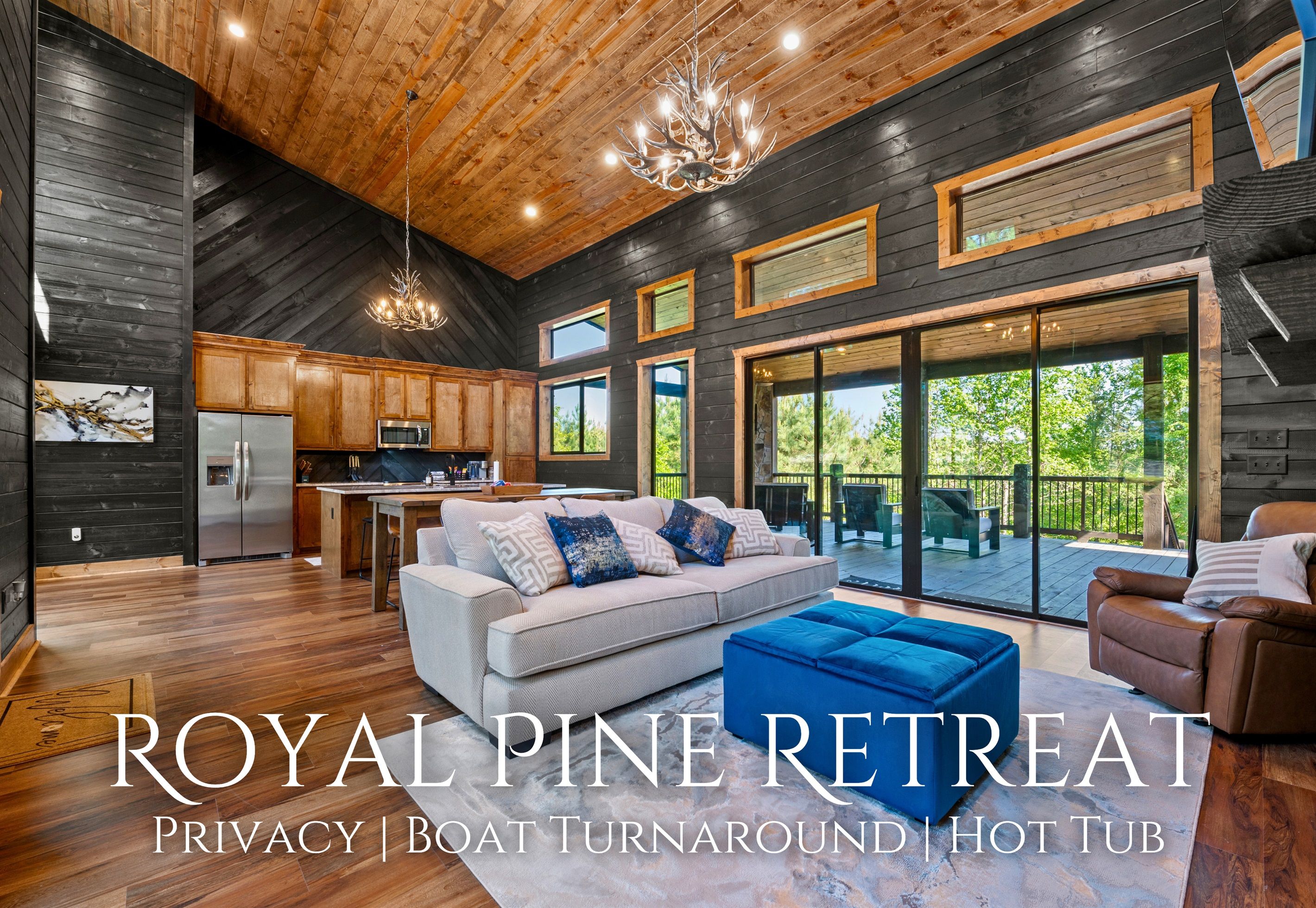 A spacious living area with a large sofa, a coffee table, and a fireplace. There is a kitchen area with modern appliances and wooden cabinets. The room has large windows offering a view of the outdoors.