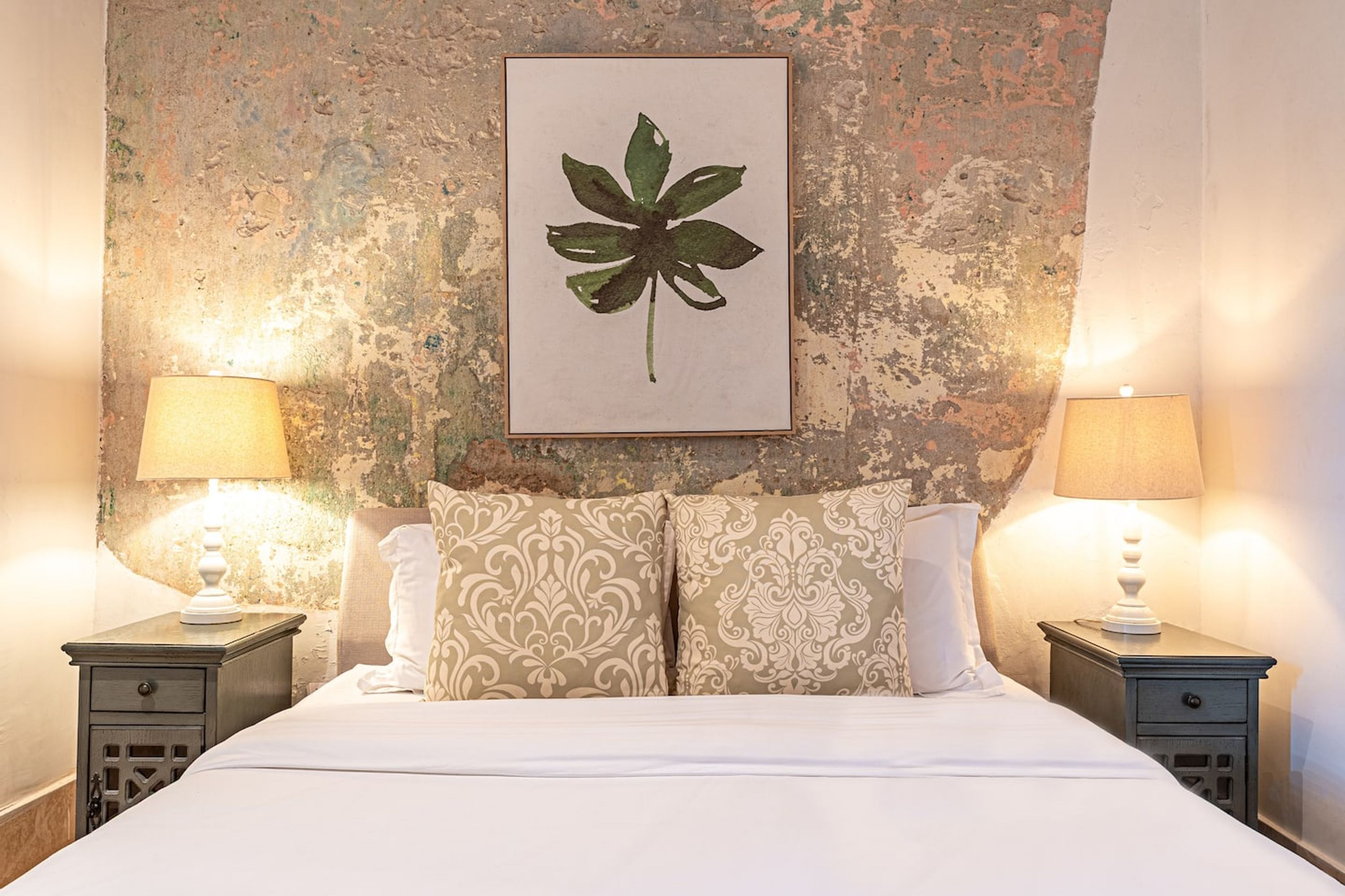 Una cama con una colcha blanca, dos mesitas de noche con lámparas, y una pared con textura y una obra de arte con una hoja enmarcada.