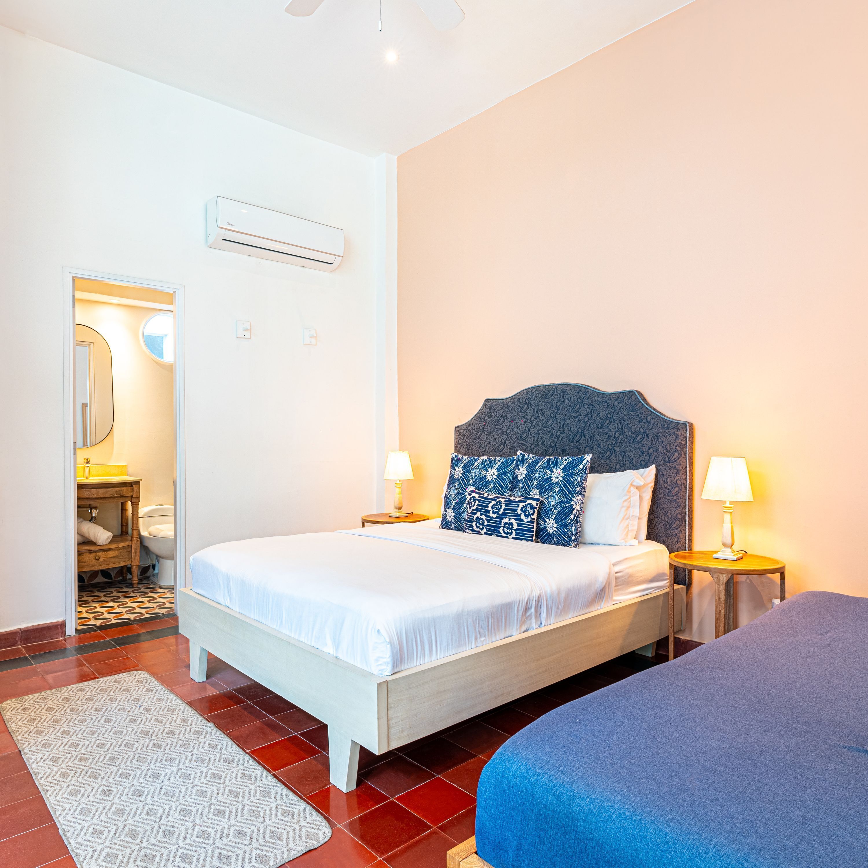 A hotel room with a bed, bedside tables, a lamp, a mirror, and a bathroom with a toilet and sink.