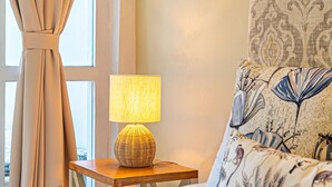 Een net opgemaakt bed met bloemenkussens, een nachtkastje met een lamp, en een raam met gordijnen.