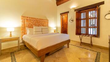 A bedroom with a wooden bed, two bedside tables, a patterned headboard, a window with wooden shutters, and a tiled floor.