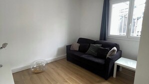 Une pièce avec un plancher en bois, un canapé de couleur foncée agrémenté de coussins, une petite table d’appoint blanche et une fenêtre avec un rideau.