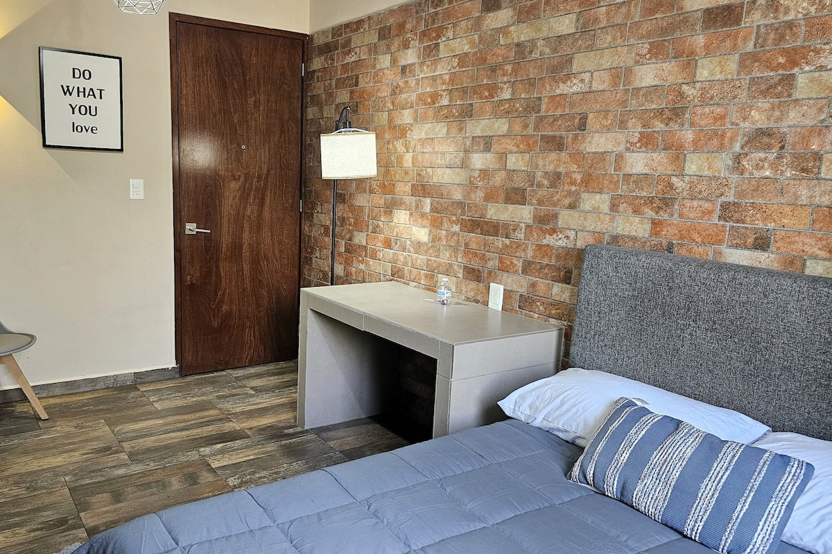 A bedroom with a brick wall, a wooden door, a desk with a lamp, a chair, and a bed with pillows.