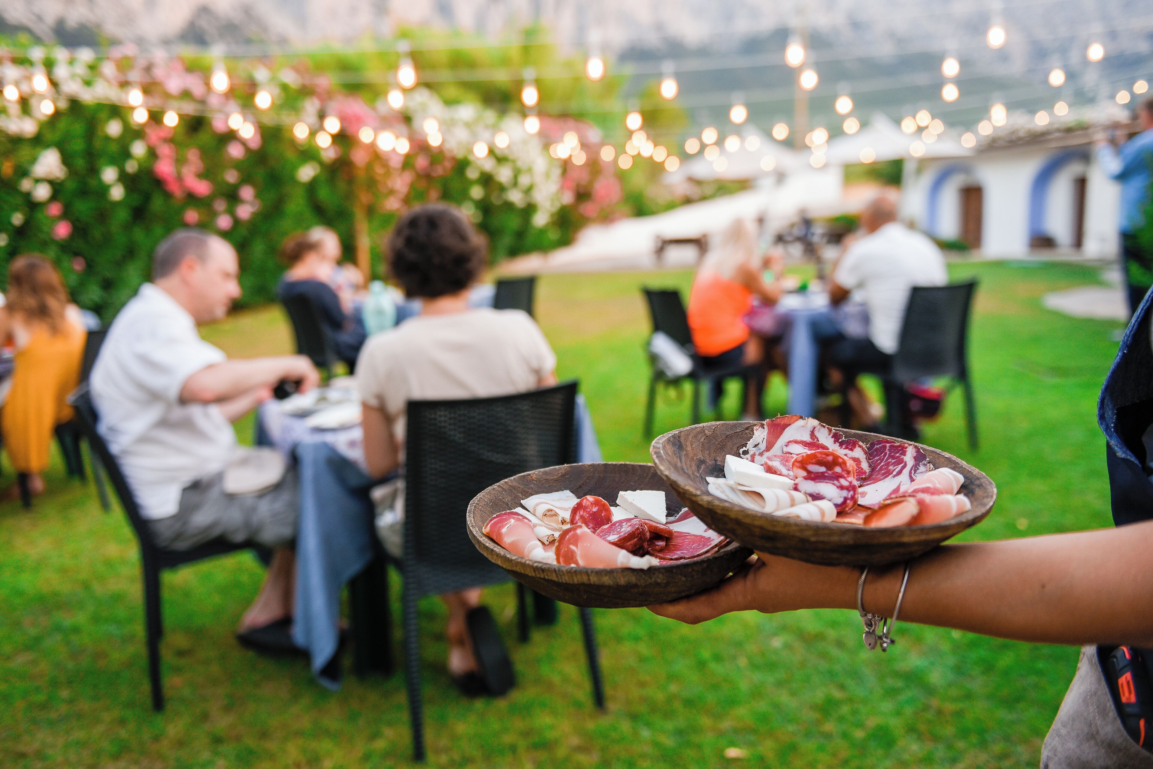 An outdoor dining area with guests seated at tables, surrounded by greenery and string lights.
