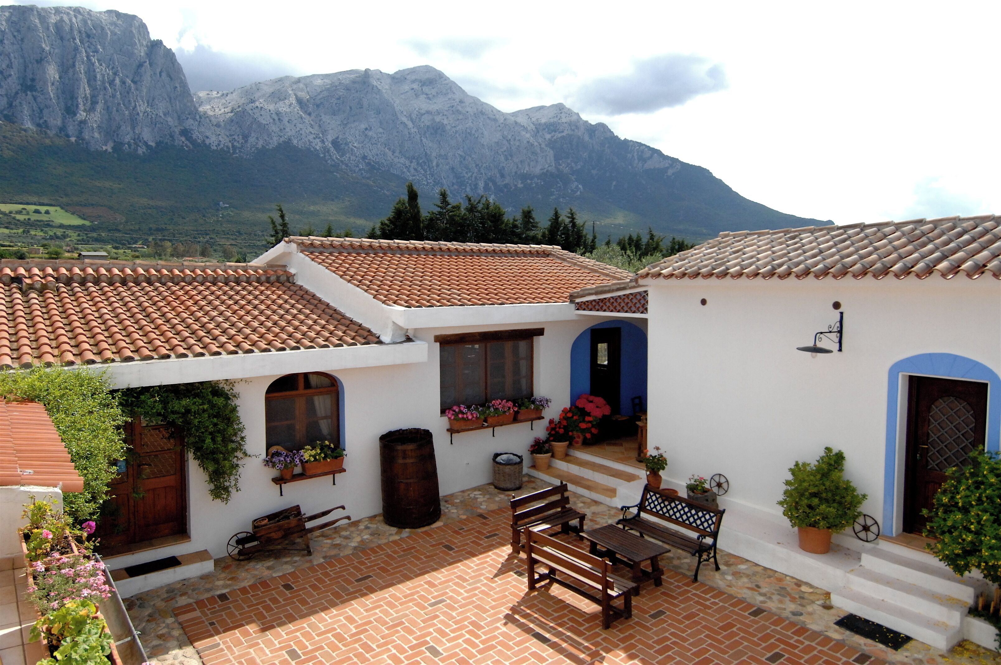 A courtyard with a tiled floor, wooden furniture, and potted plants. A mountain range is visible in the background.