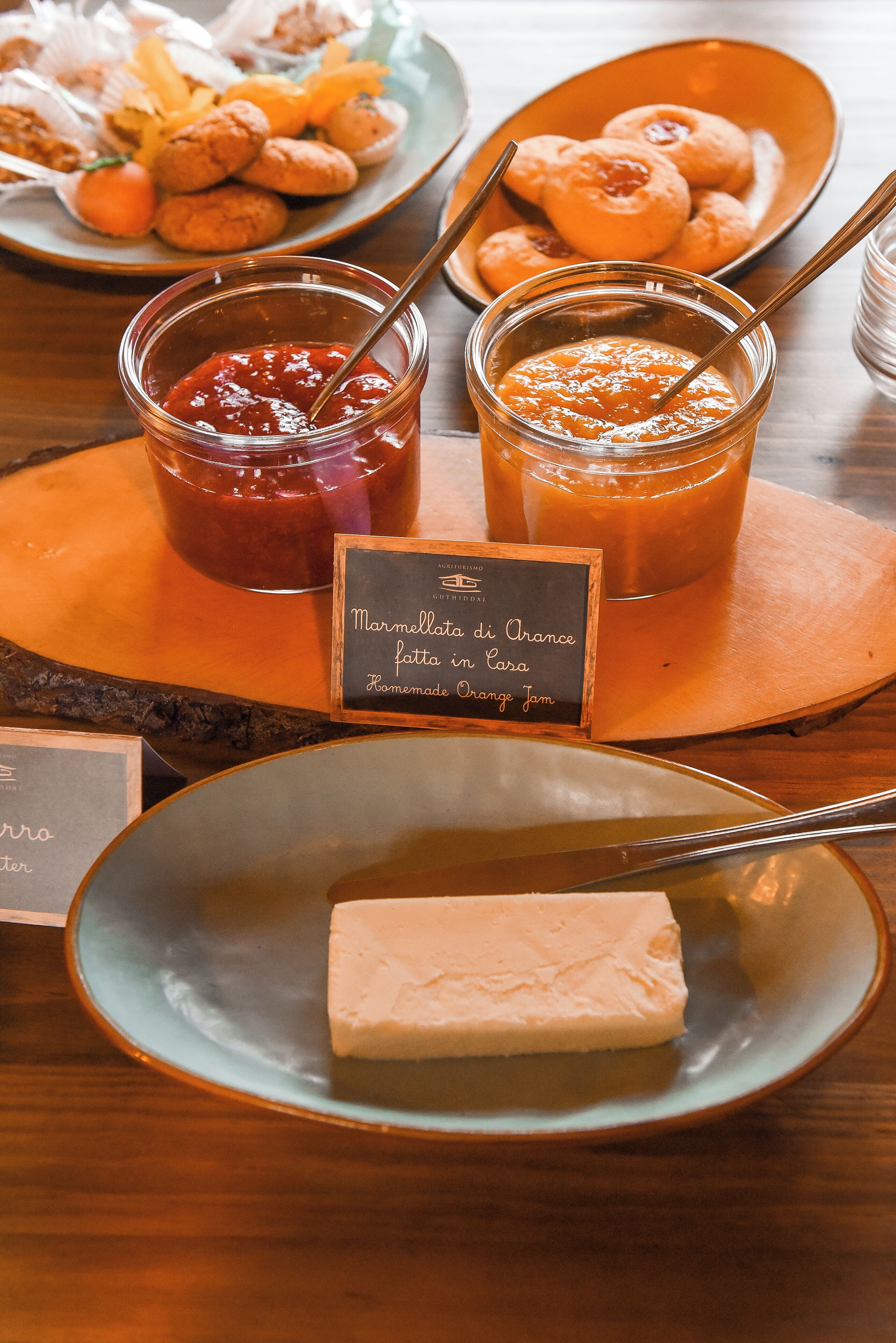 Une table présente divers aliments, notamment une assiette de fromage, des pots de marmelade et un panneau indiquant qu’il s’agit de confiture d’orange faite maison.