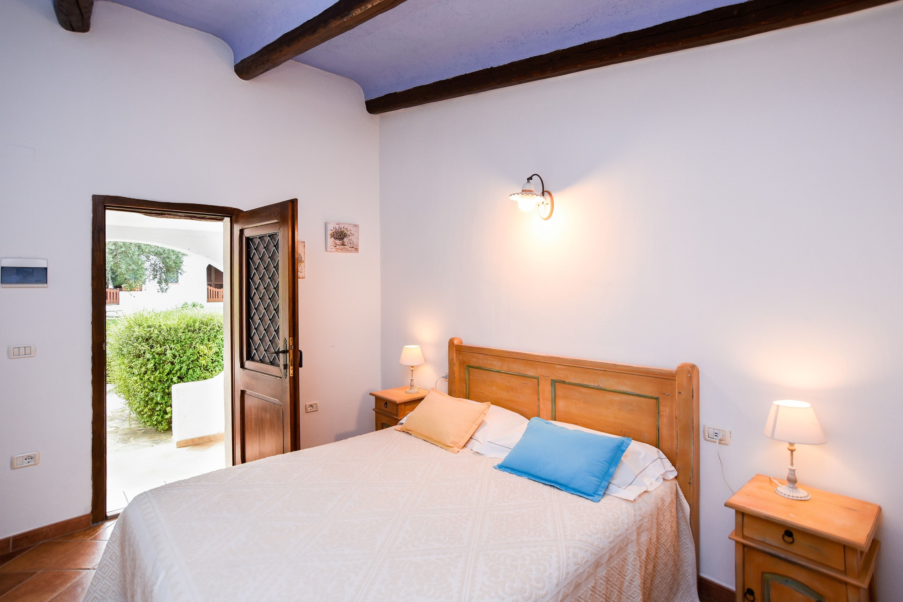 Un dormitorio con una cama de madera, dos mesitas de noche con lámparas, una puerta que da a un balcón y un aplique en la pared.