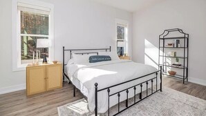 A bedroom with a bed, bedside tables, a rattan cabinet, a bookshelf, and a window with a view of trees. - Jewel of the Hidden Cove (Littleton)