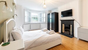A bedroom with a bed, a fireplace, a television, and a window with shutters. - Cosy and Modern Battersea Home (London)