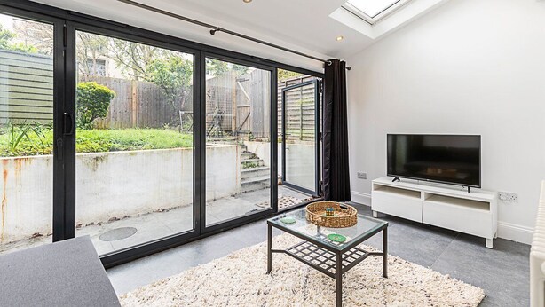 A modern living room with a large sliding glass door leading to a backyard, a flat-screen TV on a white stand, and a glass coffee table with a woven basket. - Cosy and Modern Battersea Home (London)