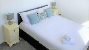 Una cama bien hecha con ropa blanca y una toalla doblada, dos mesitas de noche con lámparas y un suelo alfombrado.