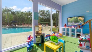 Une chambre dotée d’une grande fenêtre donnant sur une piscine, d’une télévision et d’un espace de jeux pour enfants avec des jouets et une petite table.