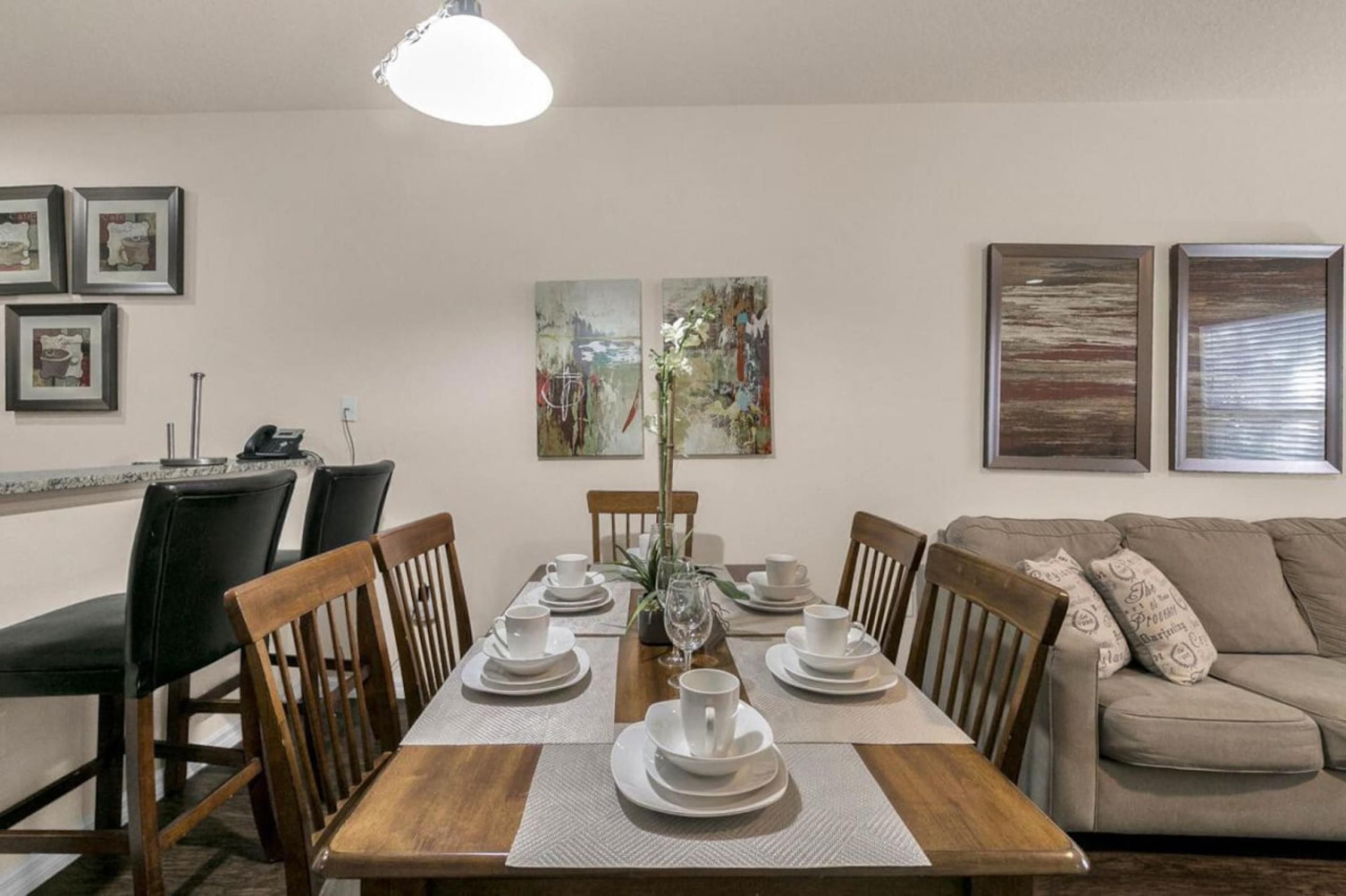 A dining area with a wooden table set for four, surrounded by chairs, with artwork on the walls and a sofa in the background.