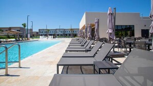 A poolside area with lounge chairs and umbrellas, a clear blue pool, and a modern building in the background. - Luxury 3bdr House Near Disney 7527 (Kissimmee)
