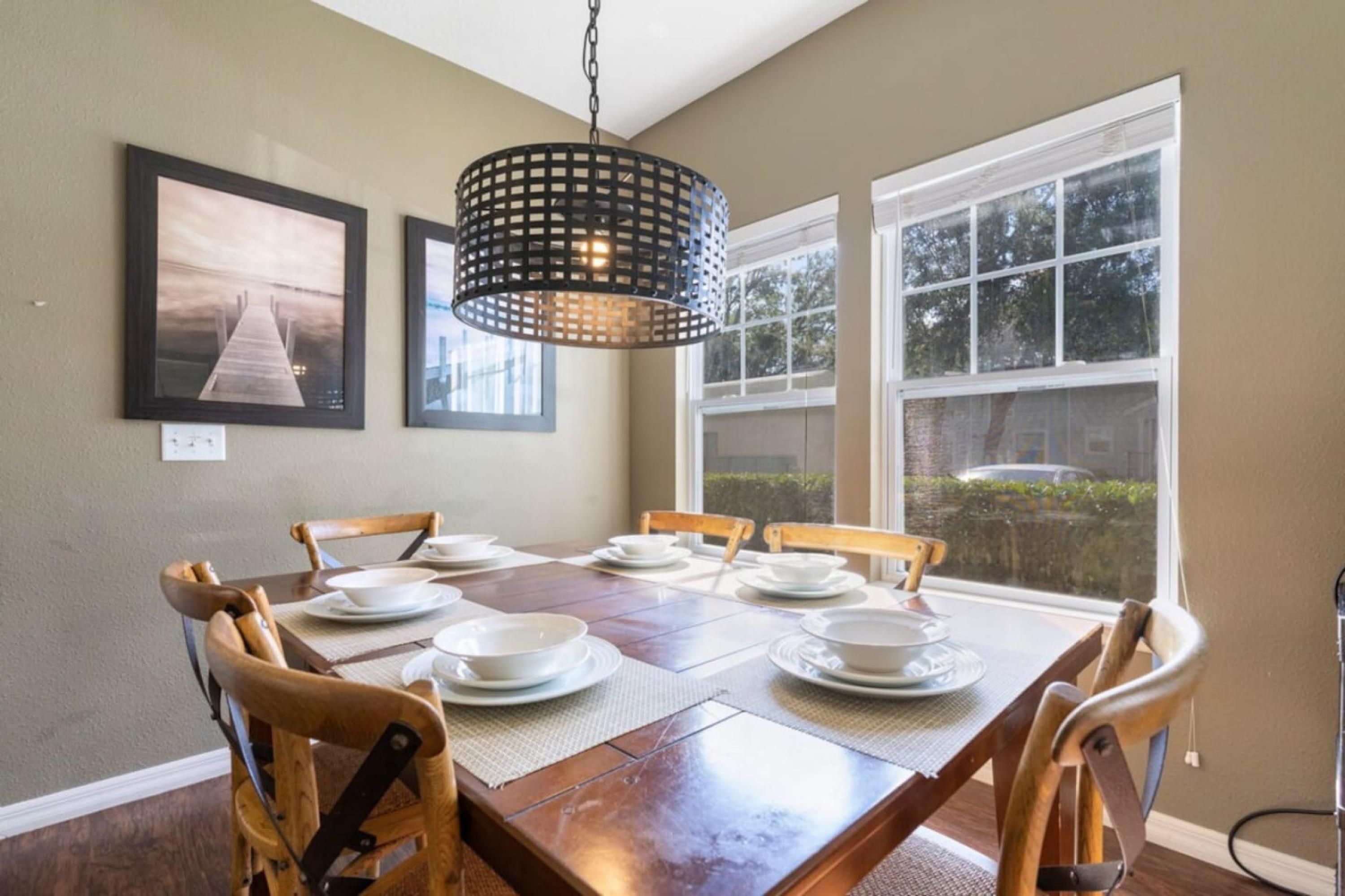 A dining area with a wooden table set for four, surrounded by chairs, with a hanging light fixture above and a window with a view of greenery.
