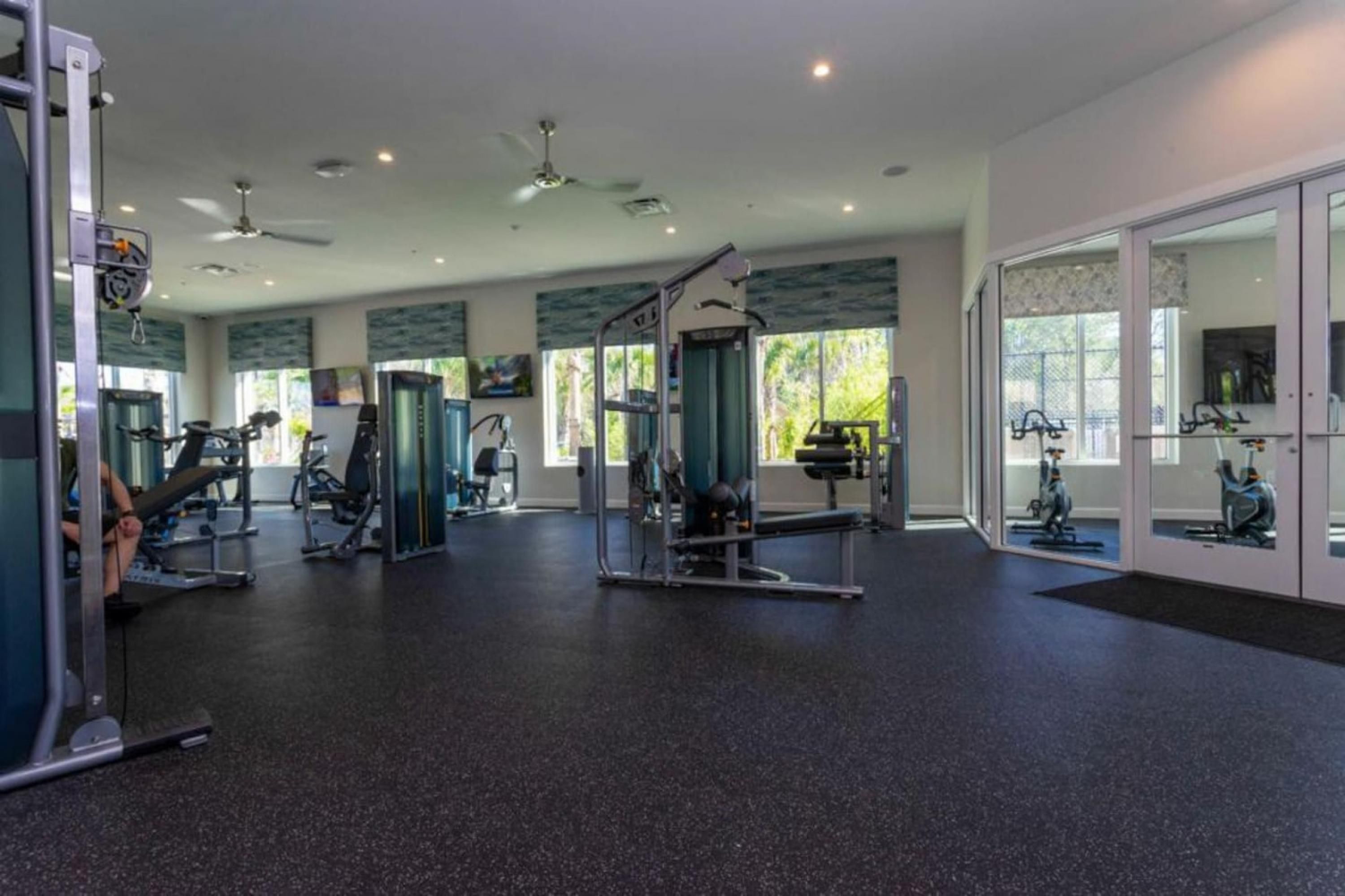 Un gimnasio amplio con diverso equipo de ejercicio, ventanas grandes y un ventilador de techo.