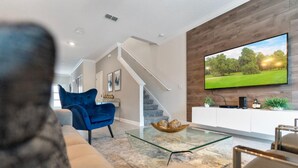 A modern living room with a glass coffee table, a blue armchair, a sofa, and a TV mounted on a wooden wall. - Amazing 5bdr Disney Vacation With Pool - 4365 (Kissimmee)