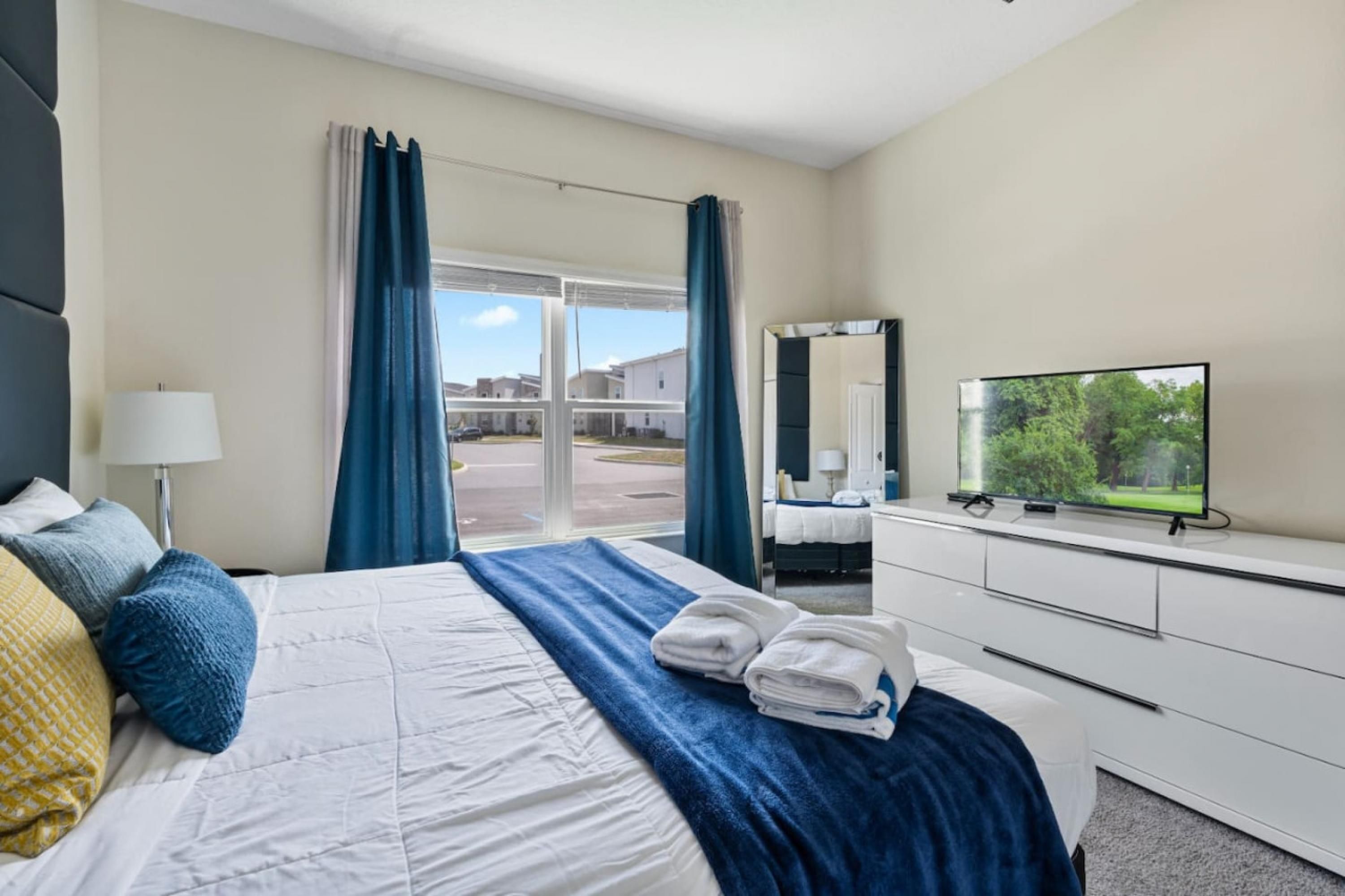 A hotel room with a large bed, a TV, a dresser, and a view of a residential area through the window.