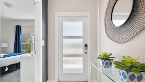 A hotel room with a white door, a bed with a blue bedspread, a window with a blue curtain, and two potted plants on a glass shelf. - Amazing 5bdr Disney Vacation With Pool - 4365 (Kissimmee)