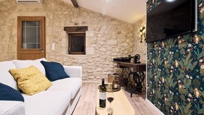 A cozy living room with a stone wall, a white sofa with blue cushions, a wooden coffee table with a bottle of wine and a glass, and a television mounted on the wall. - Gîte au Cœur des Vignes (Montagne)