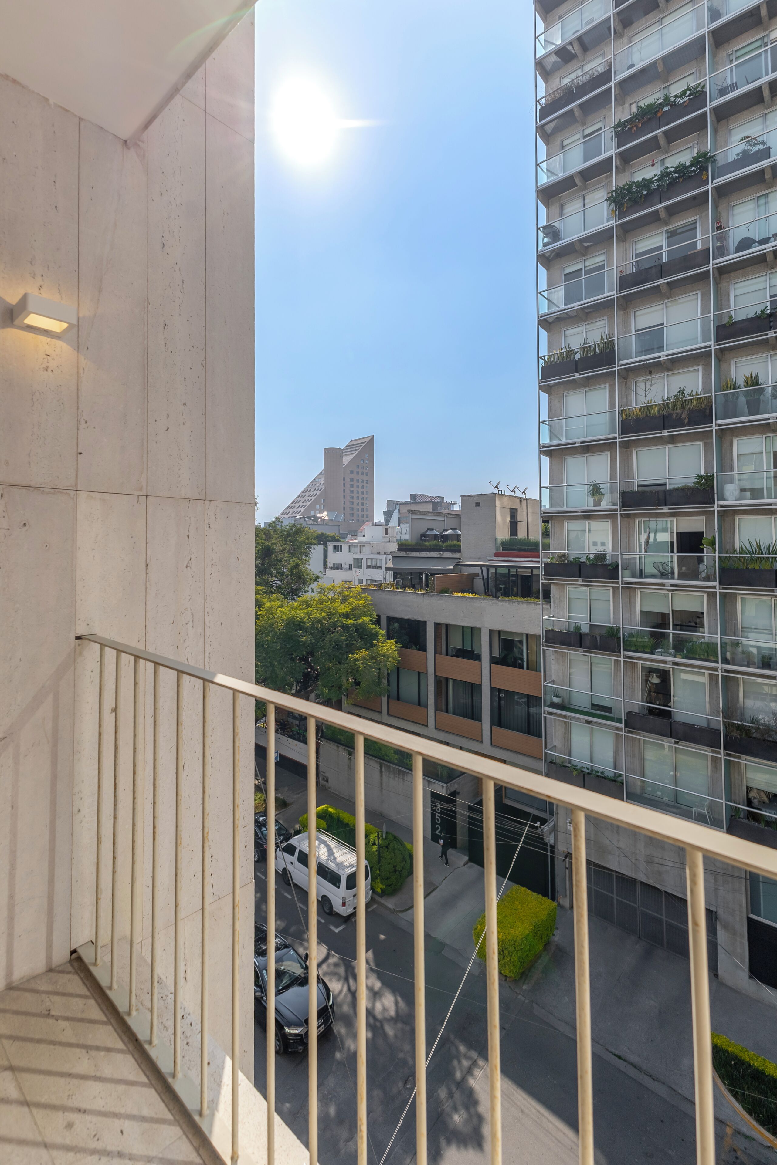 A balcony with a view of a cityscape, including a tall building with balconies and a street with parked cars.