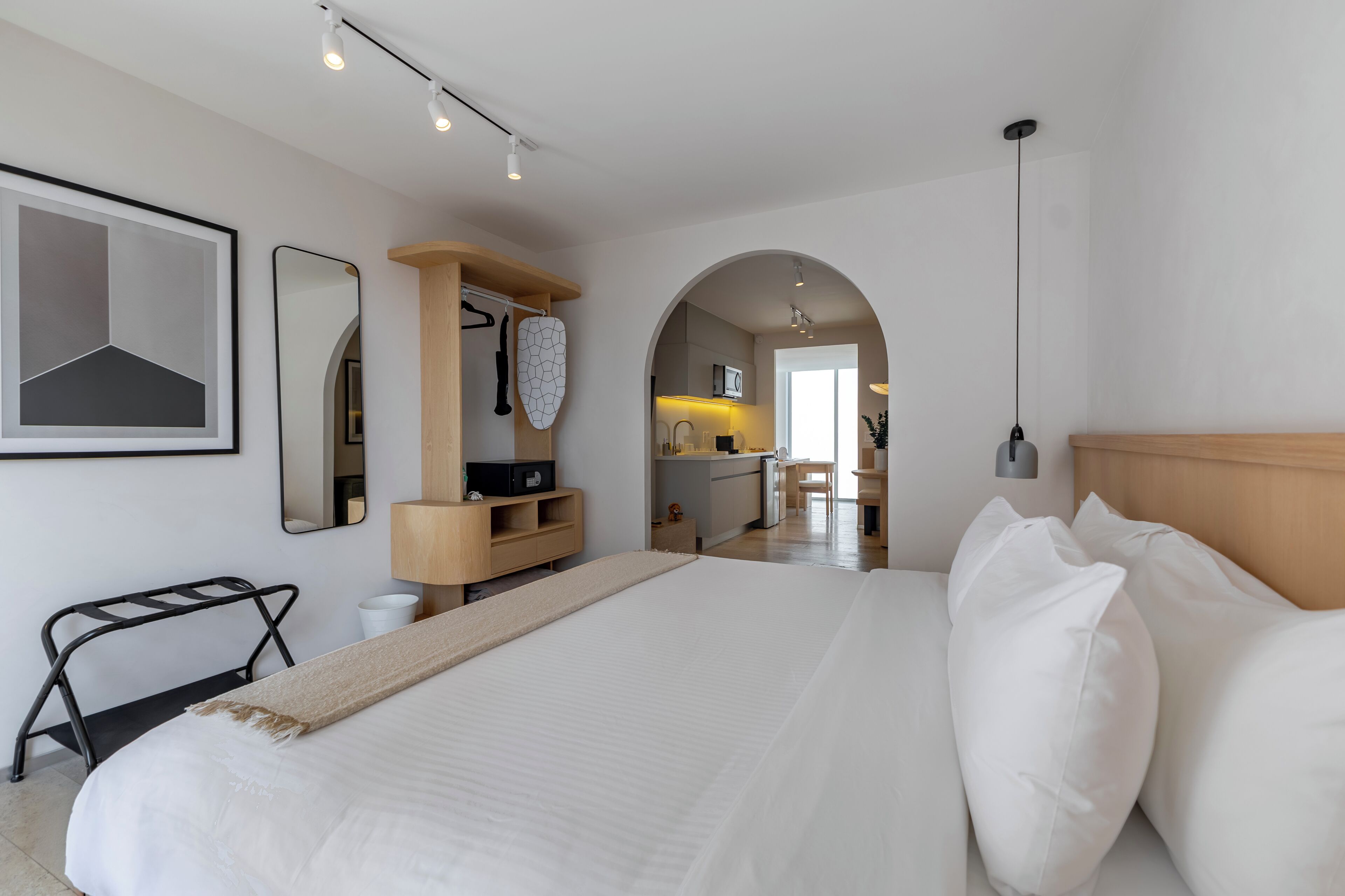 A modern bedroom with a large bed, a wooden headboard, a bedside table, a mirror, and an arched doorway leading to a kitchen area.