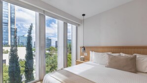A modern hotel room with a large bed, a wooden headboard, a nightstand with a lamp, and a view of the city through large windows.