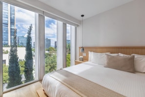 A modern hotel room with a large bed, a wooden headboard, a nightstand with a lamp, and a view of the city through large windows.
